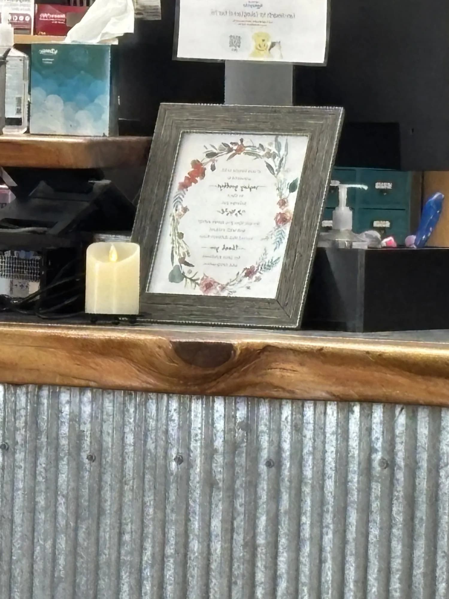 A lit flameless candle on a rustic wood counter next to a framed watercolor wreath, a box of tissues, and hand sanitizer at a veterinary clinic