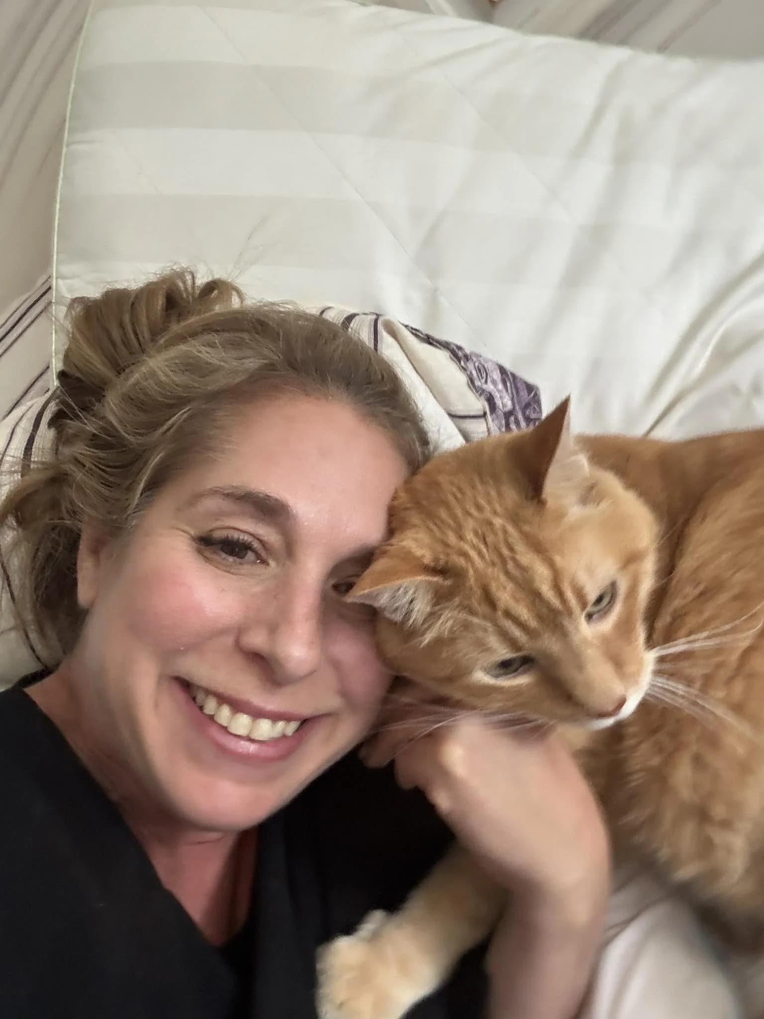 Laura lying on a pillow with Titus, her orange tabby cat, resting his head against her hand
